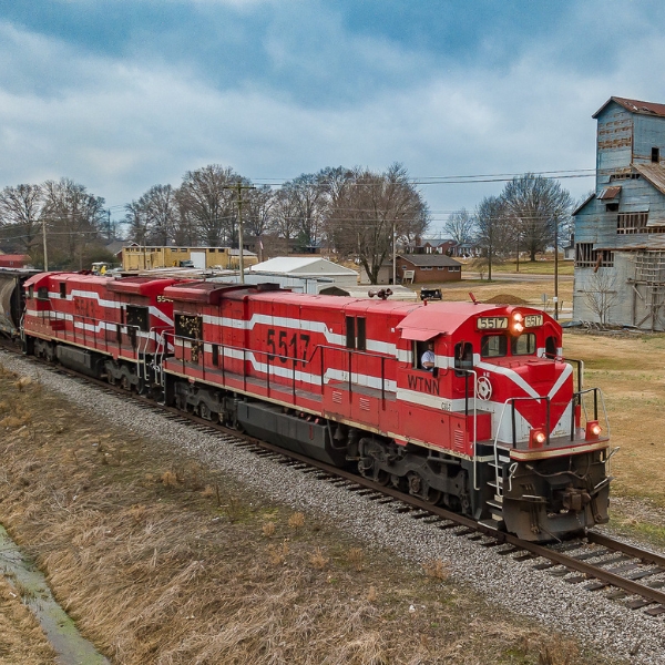red train on railroad track with old barn and houses behind